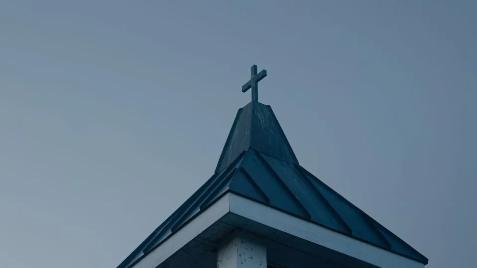 A church bell tower topped with a cross against an overcast evening sky